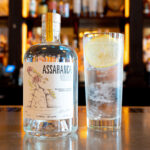 A bottle of Assaranca Vodka sits on a polished bar top next to a tall glass filled with ice, a clear mixer, and a fresh lemon wheel. The bottle features a white label with an illustration of a rowan tree and text indicating it is produced in Donegal, Ireland. The background is a softly blurred view of a backbar with various bottles on shelves and warm, ambient lighting.