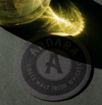 A circular Ardara Single Malt Irish Whiskey coaster sits on a gray surface, partially in shadow. A bright, golden light refraction from a nearby glass vessel dances across the top of the frame and spills onto the coaster, highlighting its dark texture and the "A" logo in the center.