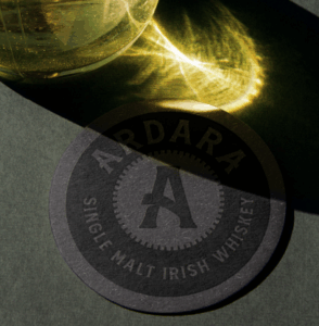 A circular Ardara Single Malt Irish Whiskey coaster sits on a gray surface, partially in shadow. A bright, golden light refraction from a nearby glass vessel dances across the top of the frame and spills onto the coaster, highlighting its dark texture and the "A" logo in the center.