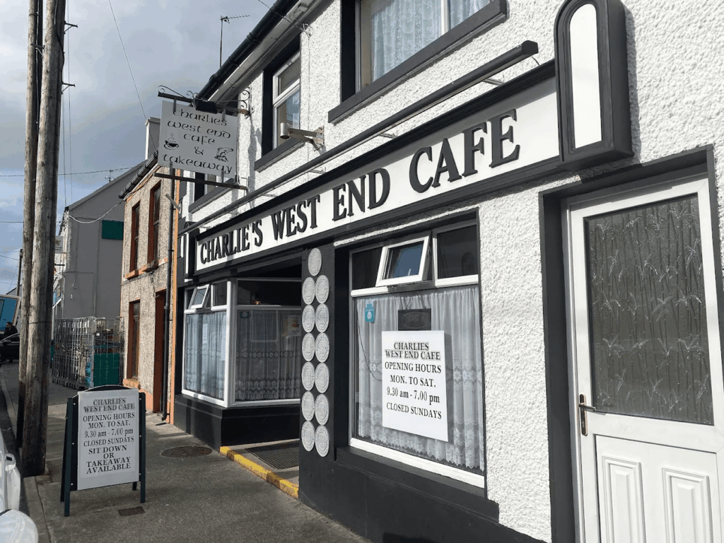 A side-angle street view of the exterior of "MOLloy's WEST END CAFE," a two-story white building with black trim. A small white sign board sits on the sidewalk in front of the entrance under a cloudy sky.