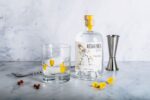 A bottle of Assaranca Vodka is positioned on a marble surface next to a glass filled with a clear liquid and a large ice cube. The scene is accented with bright yellow flower petals scattered around the base of the bottle and inside the glass, alongside a silver jigger and a long bar spoon in the background.