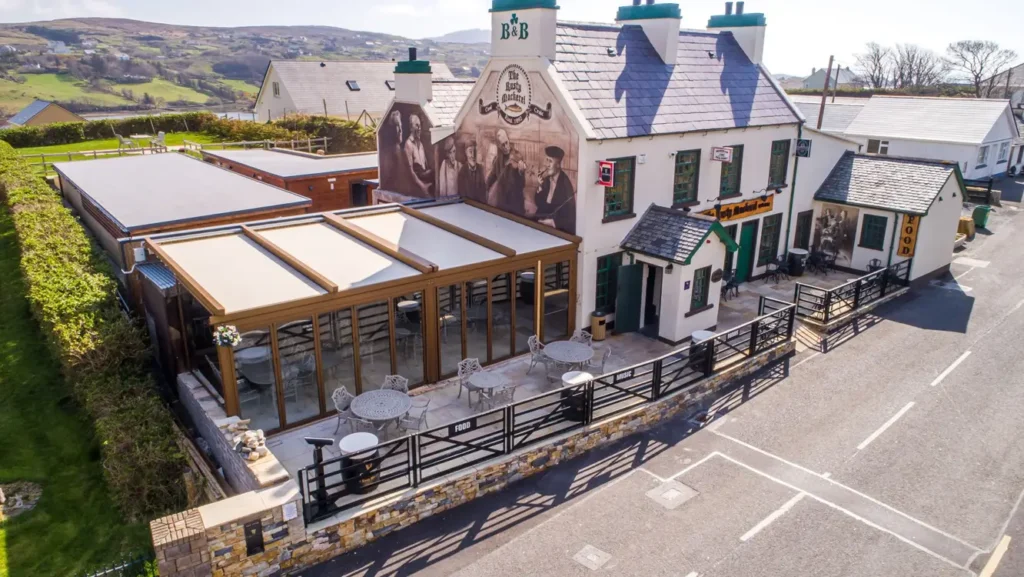 An aerial view of a traditional two-story building featuring a large mural of a seated group on its side wall. The building has a modern outdoor patio area with a retractable pergola and multiple tables, all enclosed by a low stone wall next to a paved road.