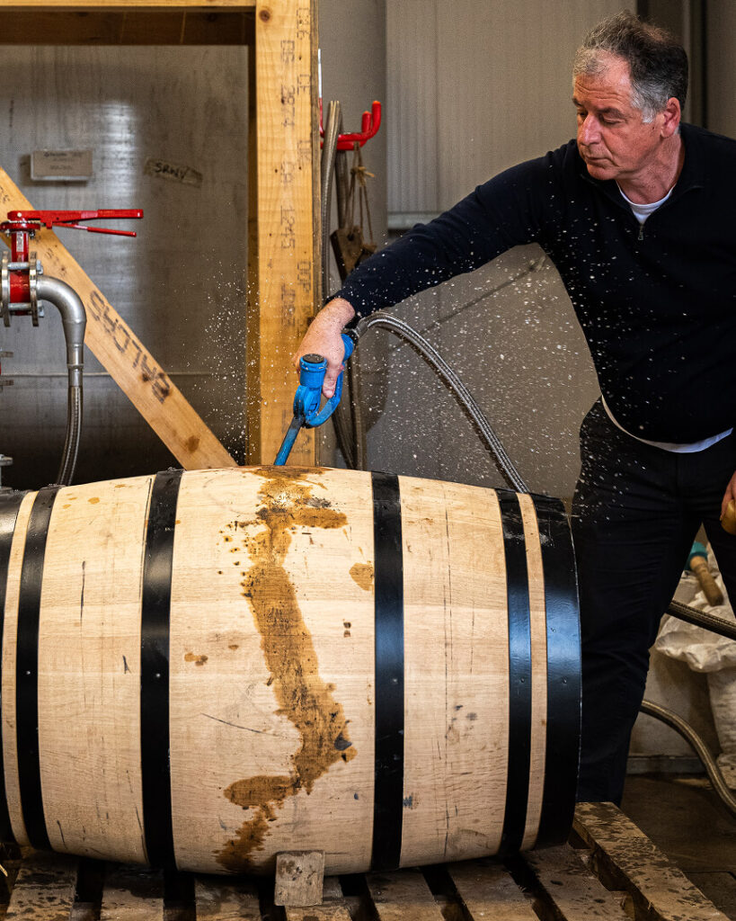 A man in a black long-sleeved shirt uses a blue hose to spray liquid onto a light-colored wooden whiskey barrel with black metal hoops.