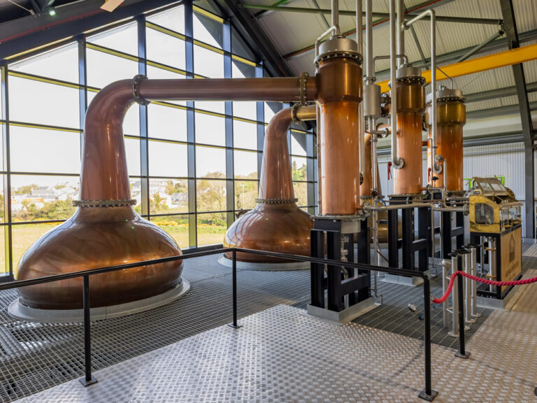 Three large, gleaming copper pot stills connected by intricate piping, situated on a metal grate floor inside a modern distillery with a massive glass wall overlooking green fields.