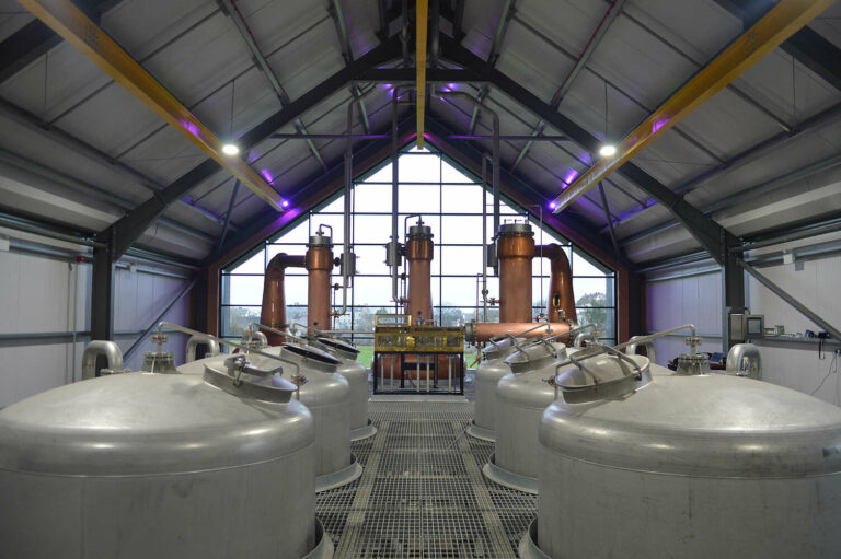 In a symmetric, wide-angle shot of the Ardara Distillery interior, two rows of large, stainless steel fermentation tanks lead the eye toward three gleaming copper pot stills in the background. The facility features a high, gabled ceiling with industrial beams and purple accent lighting, while a massive floor-to-ceiling glass wall at the far end offers a view of the landscape outside.