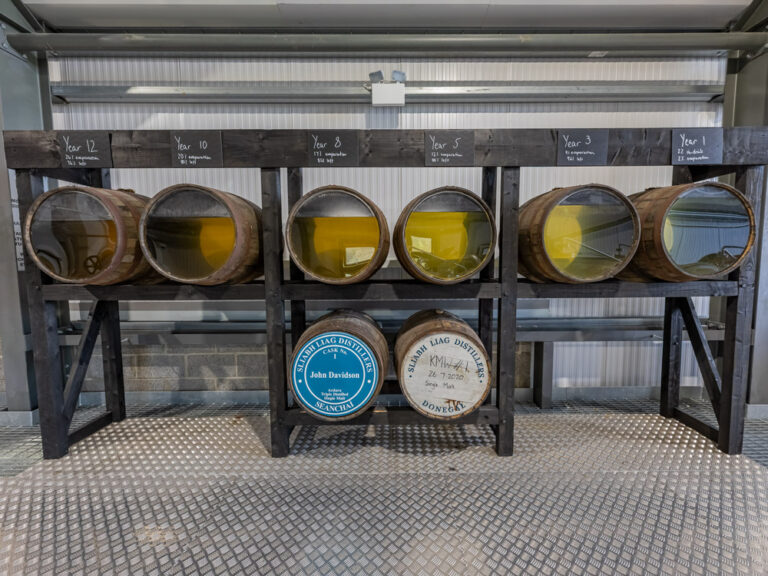 Six small wooden barrels with glass ends showing whiskey at various stages of aging, displayed on a black wooden rack labeled with years 1 through 12.