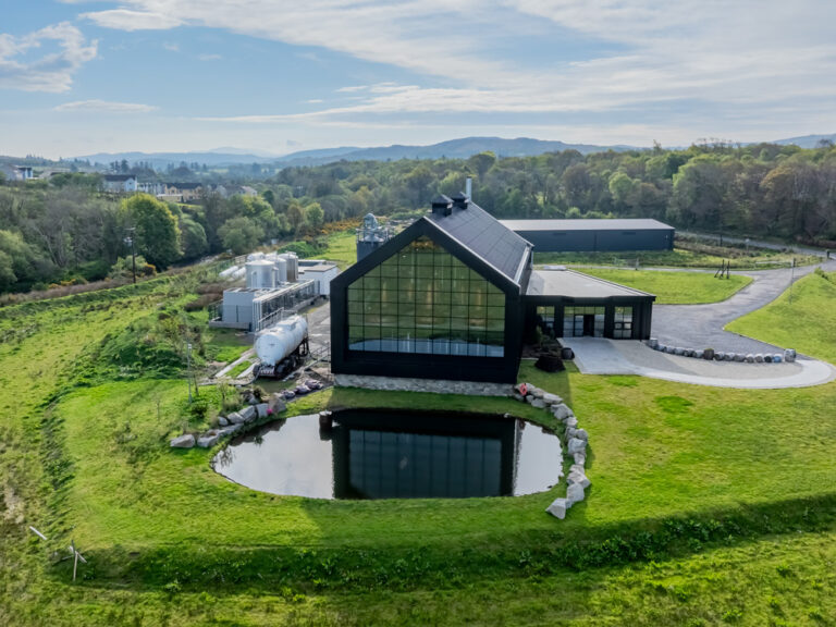 An aerial view of the modern, black-gabled Ardara Distillery building with its expansive glass facade reflecting a nearby pond, situated among lush green hills and industrial equipment.