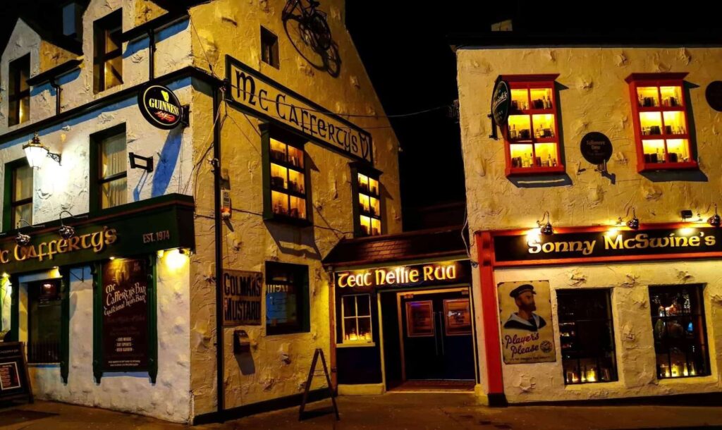 A nighttime exterior shot of a traditional white-walled Irish pub and restaurant complex. The buildings feature several storefronts, including McCafferty's and Sonny McSwine's, with windows glowing from warm interior lighting. Decorative signs, including a Guinness logo and a "Colman's Mustard" advertisement, are visible on the textured stone walls.