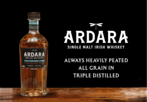 A bottle of Ardara Single Malt Irish Whiskey (2025 Edition) stands on a wooden surface against a black background. To the right of the bottle, large white text features the Ardara logo with a dragonfly icon, followed by the descriptors: "ALWAYS HEAVILY PEATED," "ALL GRAIN IN," and "TRIPLE DISTILLED." Faint wisps of smoke curl across the wooden foreground.