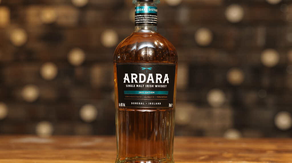 Front view of a clear glass bottle filled with amber-colored Ardara Single Malt Irish Whiskey. The black label features a teal dragonfly logo and white text. The bottle is set on a rustic wooden table with a warm, out-of-focus background.