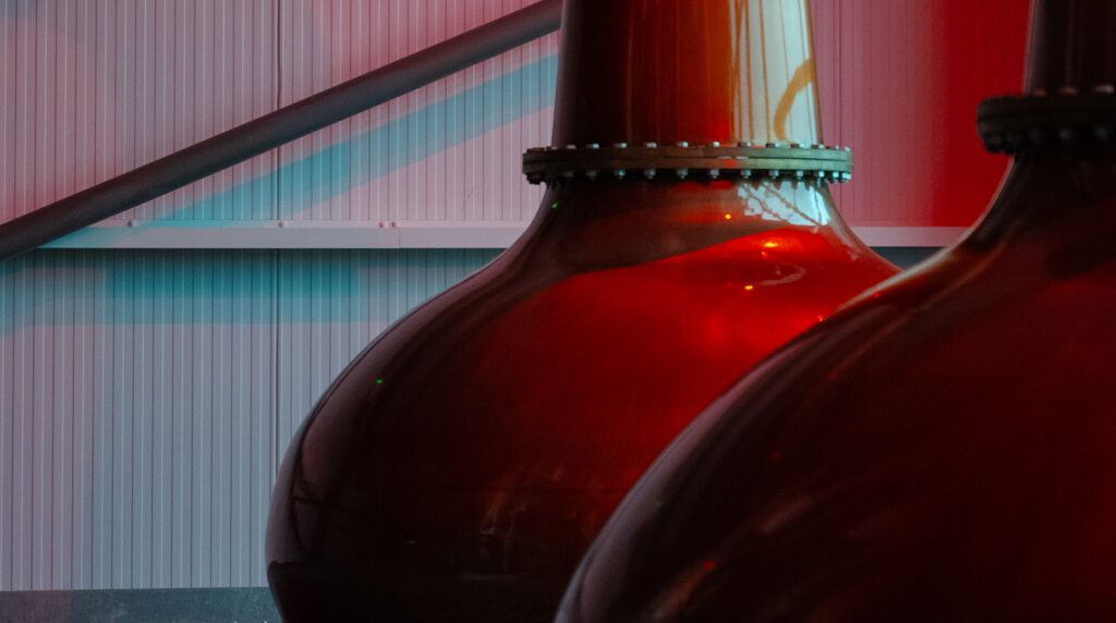 Red illuminated copper still against industrial distillery wall