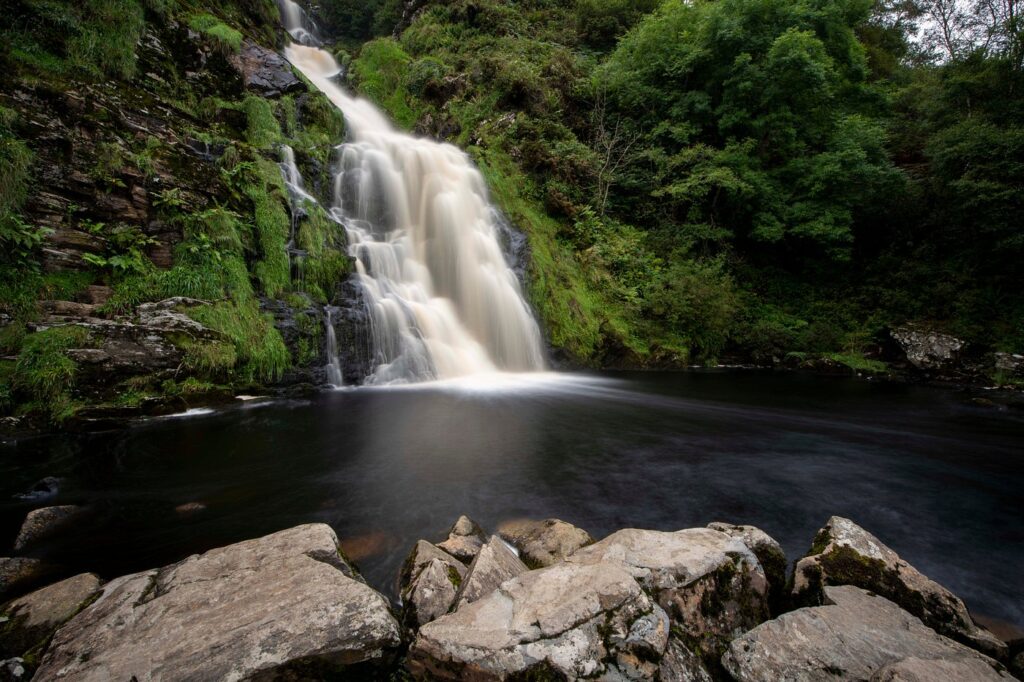 A scenic, long-exposure photograph of a waterfall cascading down a lush, green rocky cliff into a dark pool of water below. Large, jagged rocks are visible in the immediate foreground, and the dense foliage surrounding the falls creates a serene, natural atmosphere.