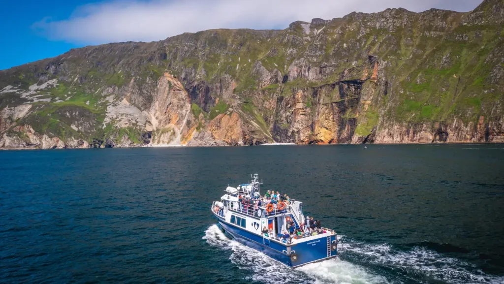 A blue tour boat filled with passengers travels across dark blue water toward the base of high, rugged green and brown cliffs under a clear sky.