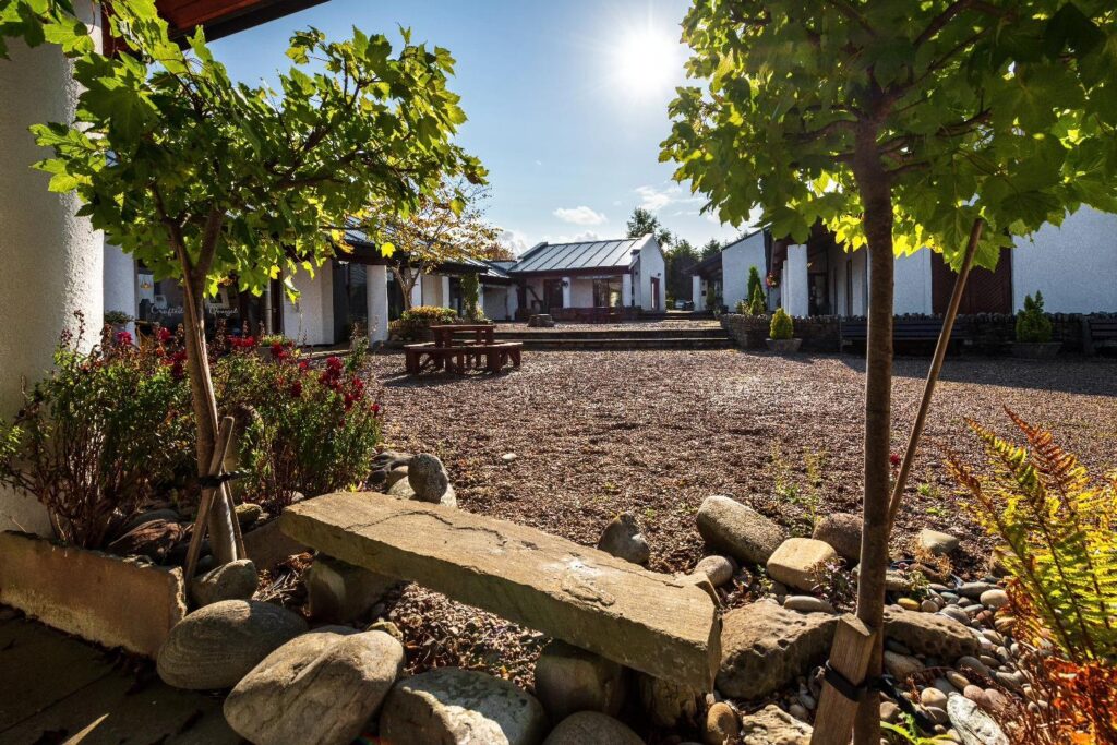 A wide-angle, sunlit view of the Ardara Distillery courtyard. In the foreground, a stone bench sits nestled among large rocks and green foliage. The background features several white, modern buildings with dark gabled roofs surrounding a central gravel area under a clear, bright sky.