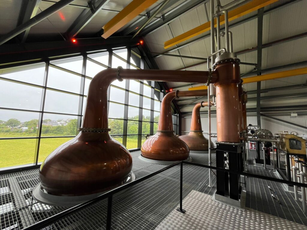 A wide-angle interior view of the Ardara Distillery production floor, featuring a row of three large, gleaming copper pot stills. The stills are positioned along a massive floor-to-ceiling glass wall that overlooks a green landscape. The facility has a modern industrial design with a high, vaulted dark ceiling, yellow structural beams, and a metallic grate floor.