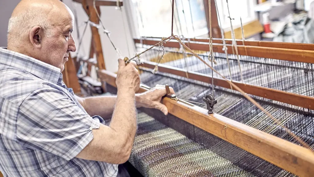 A side profile of an elderly man with a bald head and a checkered shirt operating a traditional wooden weaving loom. He is shown focused on the intricate threads of the fabric as he works indoors.