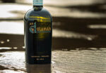 A bottle of An Dúlamán Irish Maritime Gin stands on a sandy beach at low tide. The dark glass bottle reflects the golden light of a sunset or sunrise, with wet sand and gentle ripples of water visible in the blurred foreground and background.
