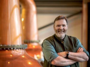 A man with graying hair and a beard smiles warmly with his arms crossed in front of two large, gleaming copper pot stills. He is wearing a green vest over a blue button-down shirt and a watch on his left wrist. The distillery equipment in the background is softly out of focus, highlighting the subject.