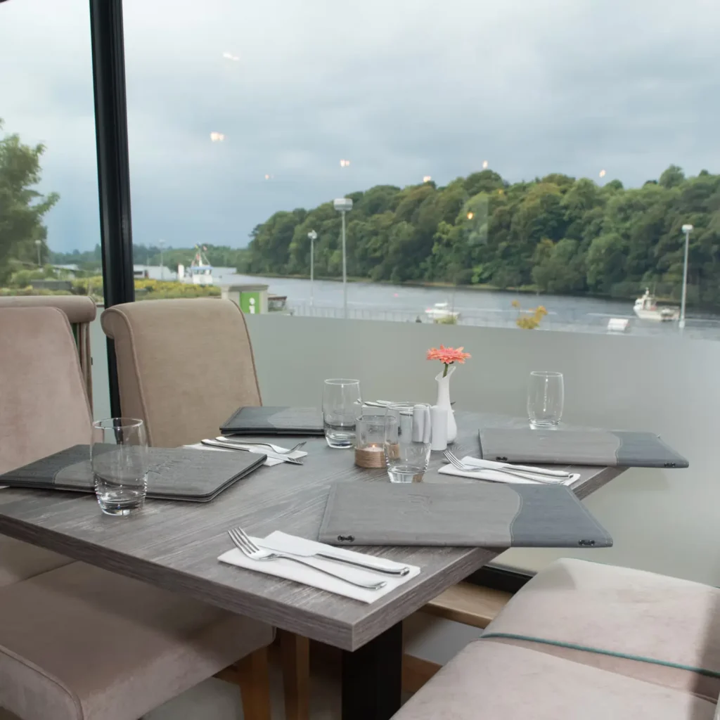 A set dining table for two in a modern restaurant with a large window offering a view of a wide river and a dense treeline on the opposite bank. The table is set with gray linens, water glasses, and a small flower in a white vase.