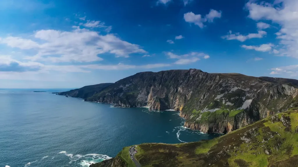 A panoramic coastal landscape featuring the massive, dark gray sea cliffs of Slieve League in Donegal, Ireland. The green and brown rugged cliffside drops steeply into the deep blue Atlantic Ocean under a bright sky with scattered white clouds. A small winding road and parking area are visible on a lower grassy ridge in the foreground.