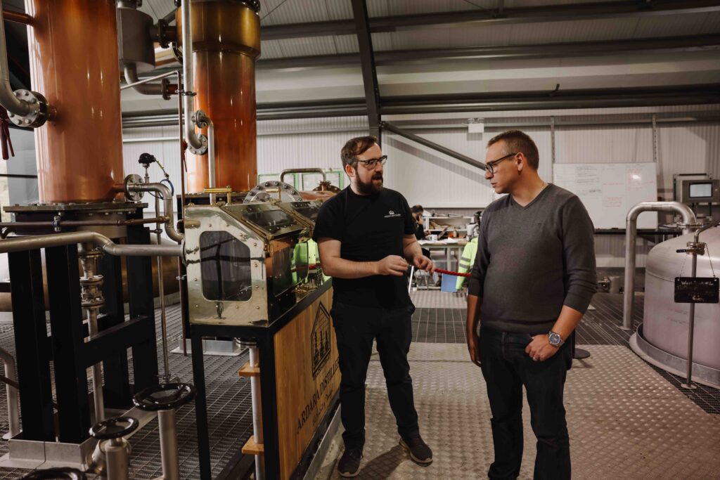 Two men in casual attire, one wearing glasses and a black t-shirt and the other a gray v-neck sweater, engaged in conversation next to copper distilling equipment and a spirit safe inside a modern industrial distillery.