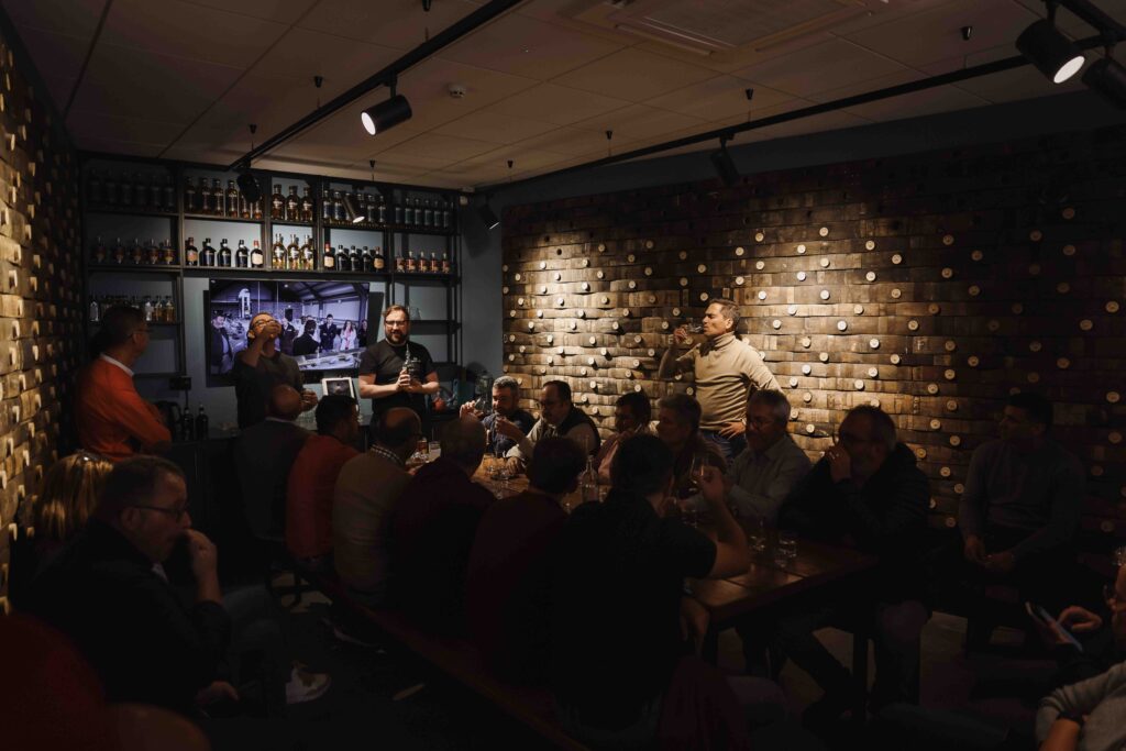In a dimly lit tasting room with walls made of stacked barrel ends, a large group of people sits around a long wooden table for a whiskey tasting session. On the left, a presenter stands near shelves of bottles and a video screen, while several attendees are captured in the act of sampling their drinks.