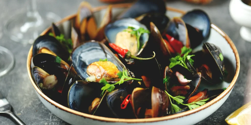A close-up shot of a bowl filled with steamed mussels in their dark shells. The dish is garnished with fresh green parsley and sliced red chili peppers, all served in a shallow white bowl with a thin brown rim.