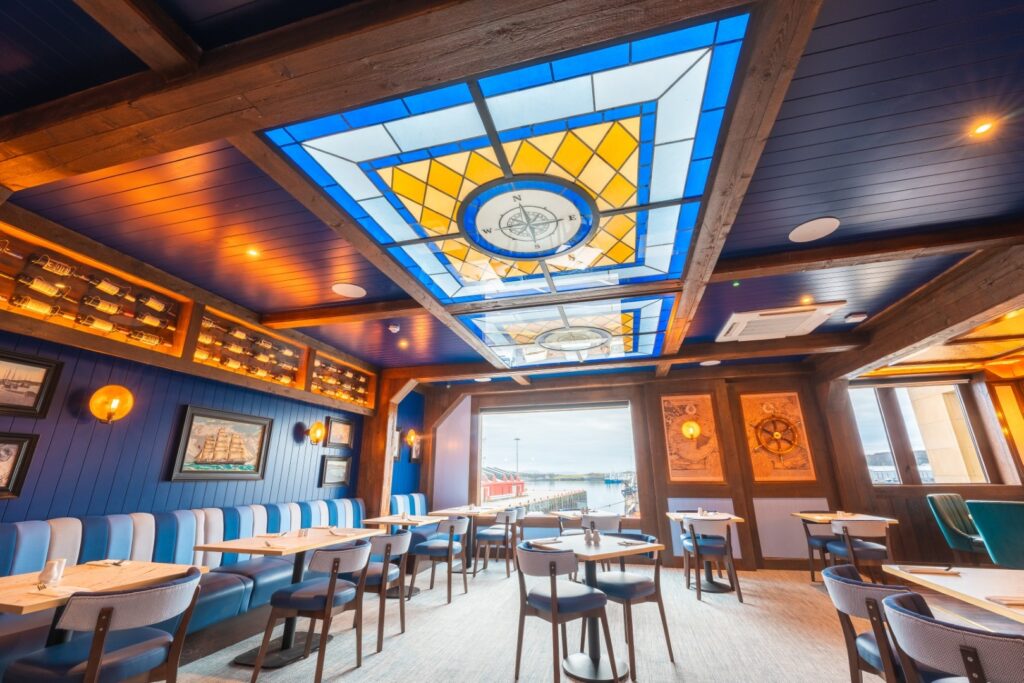 A vibrant, maritime-themed restaurant interior with blue wood-paneled walls and a large, ornate stained-glass ceiling featuring a nautical compass design in yellow and blue. The room is furnished with blue upholstered booths and modern chairs around light-wood tables, while nautical artwork and warm lighting create a cozy, coastal atmosphere.