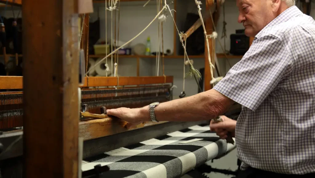 A side-profile view of a man in a checkered shirt operating a large, traditional wooden weaving loom in a workshop. He is shown focused on moving the shuttle across a black and white patterned fabric.