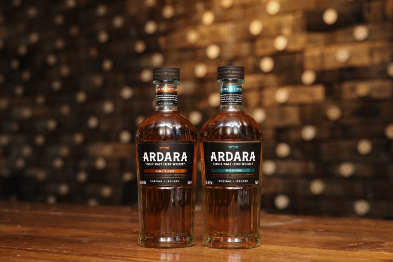 Two bottles of Ardara Single Malt Irish Whiskey sit side-by-side on a wooden surface against a backdrop of stacked barrel ends. The amber-colored spirits are presented in clear glass bottles with black labels featuring a dragonfly emblem.