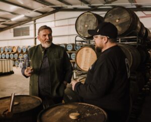 Two men holding tasting glasses and conversing in a whiskey warehouse, surrounded by stacks of aging wooden barrels.