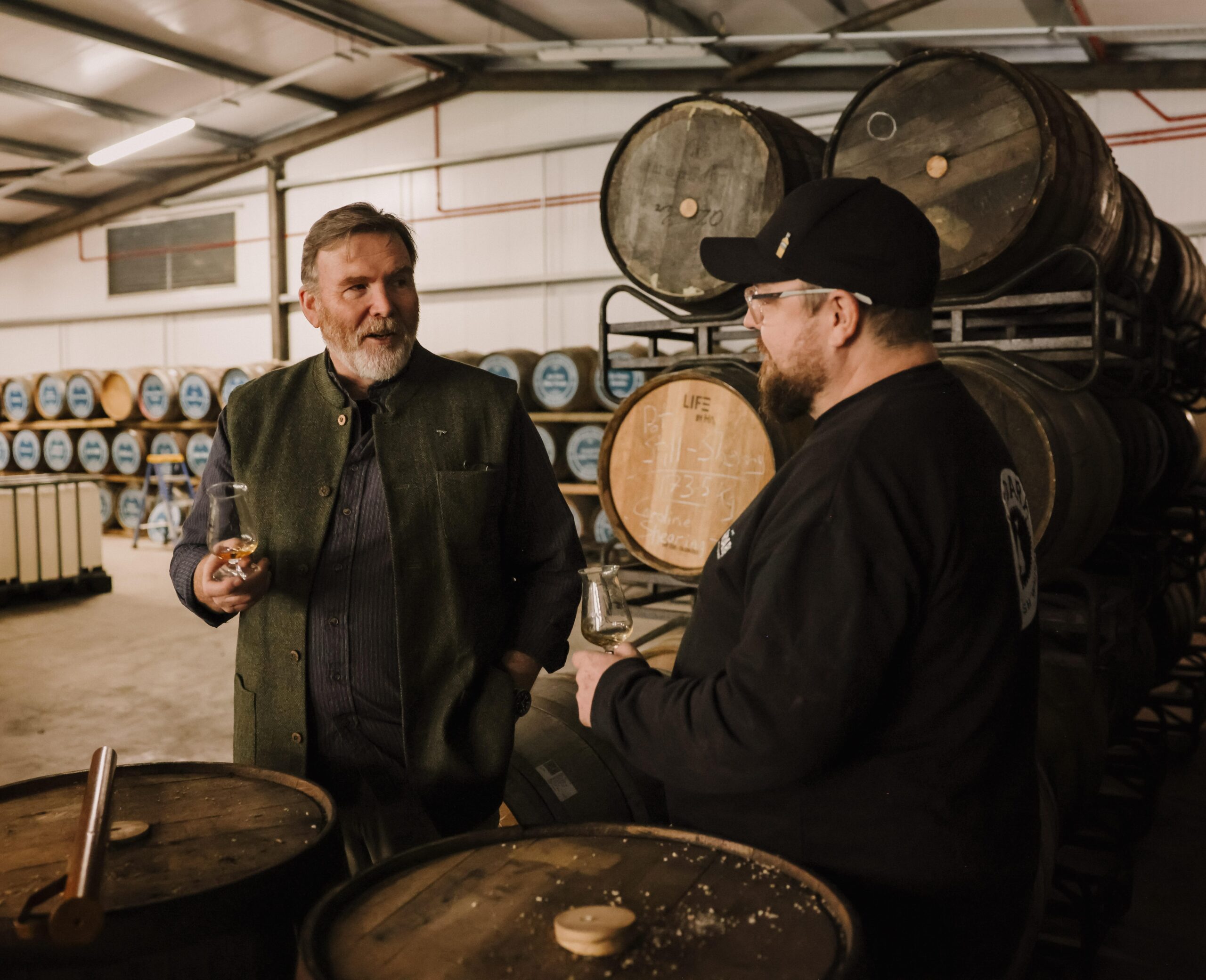 Two men holding tasting glasses and conversing in a whiskey warehouse, surrounded by stacks of aging wooden barrels.